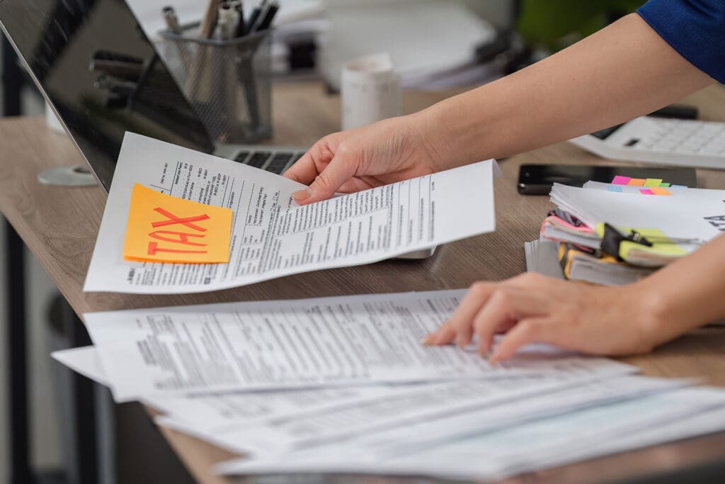 A person sorting tax paperwork on a desk with a laptop, calculator, and cell phone in the background.