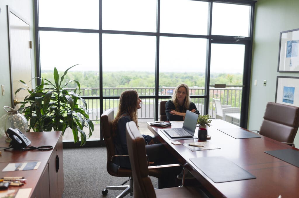 Two people sit at a long wooden desk, talking.