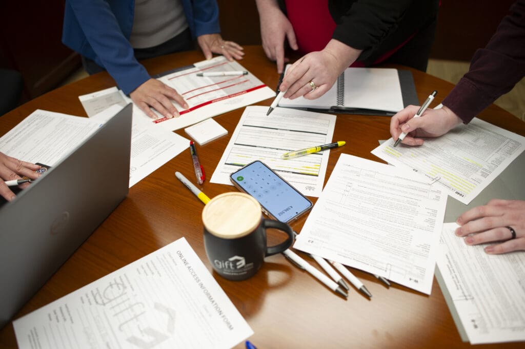The hands of a group of people working on accounting paperwork at a wooden desk.